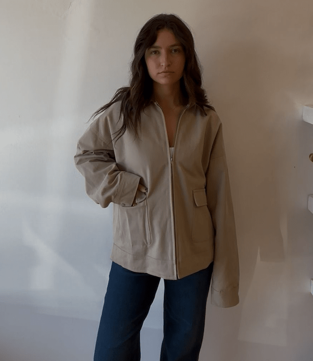 Model wearing a taupe zip-front jacket layered over a light top with dark blue jeans.