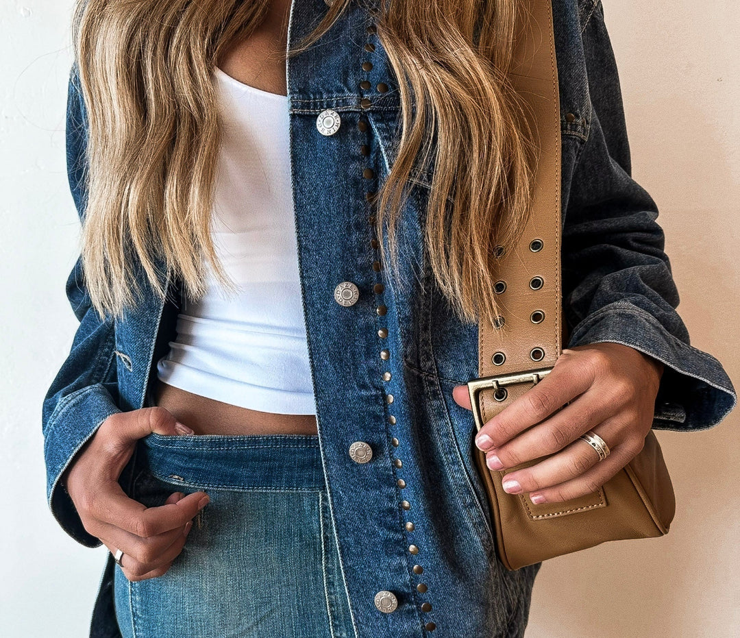 Close-up of a woman wearing a denim studded jacket layered over a white crop top and denim skirt, accessorized with a tan crossbody bag featuring a wide grommeted strap and silver rings on her fingers.