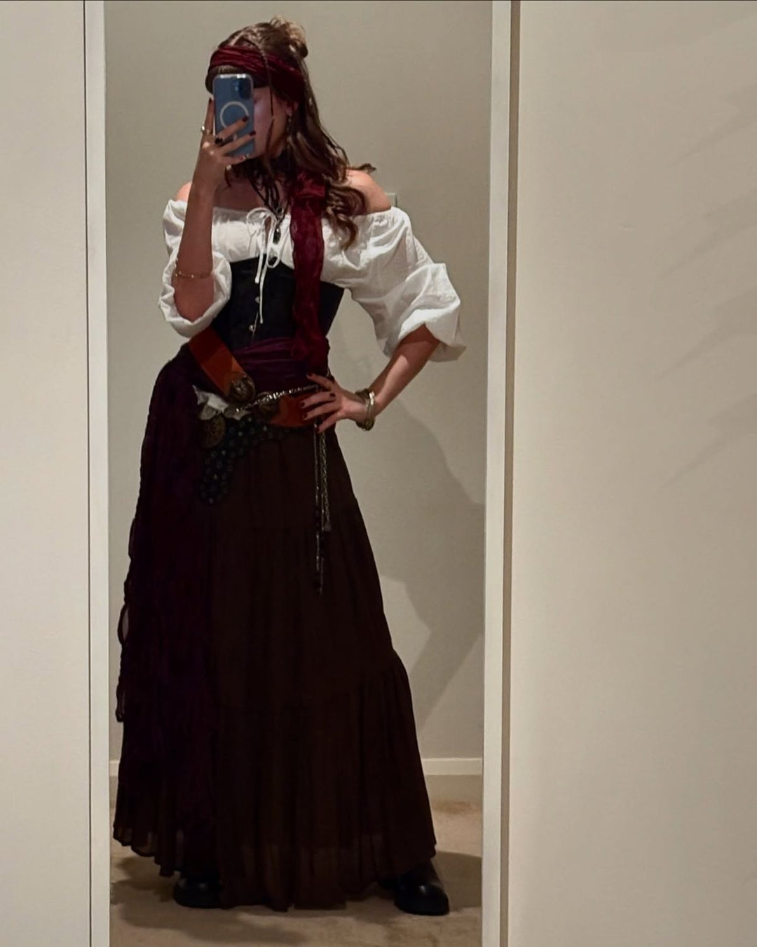A woman takes a mirror selfie dressed in a pirate-inspired costume, wearing an off-the-shoulder white blouse, black corset, long brown skirt, and layered accessories including belts, coins, and a red scarf tied around her head.
