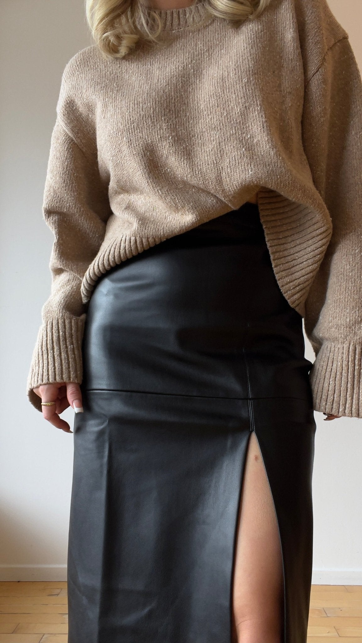 A woman wearing a soft beige knit sweater partially tucked into a sleek black leather midi skirt with a front slit, creating an effortlessly chic and modern fall outfit.