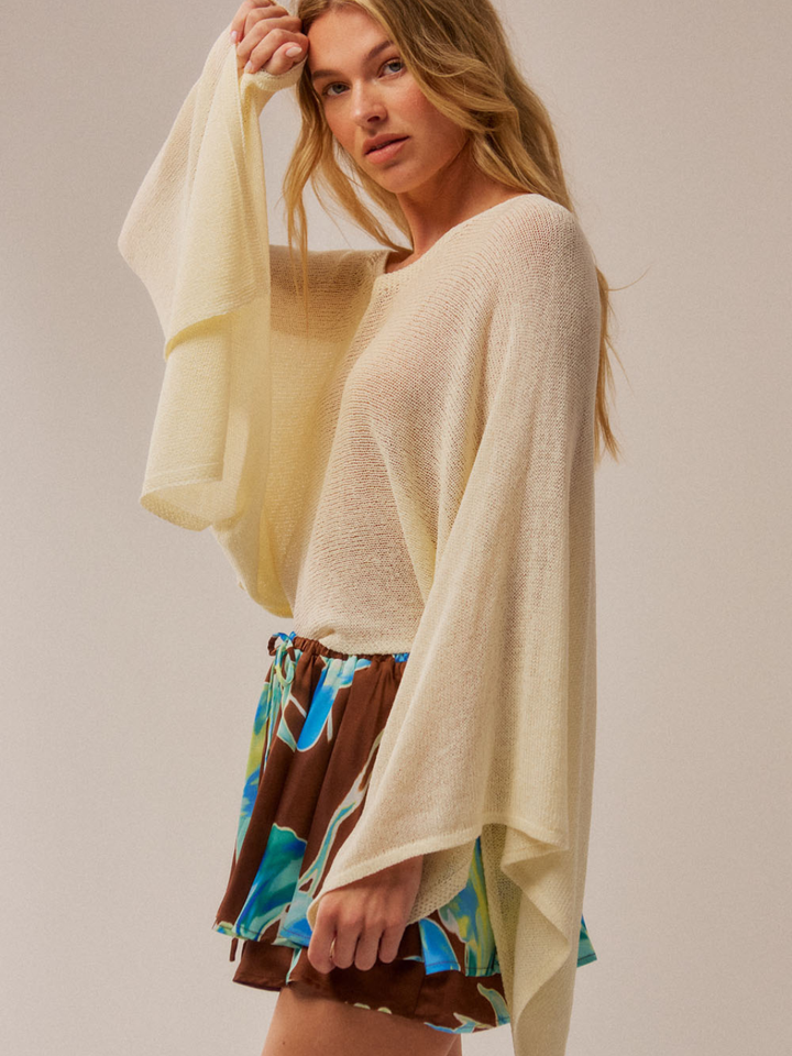 Cream Daydrift Sheer Knit Poncho Sweater worn by a woman; oversized, airy drape over a colorful skirt.