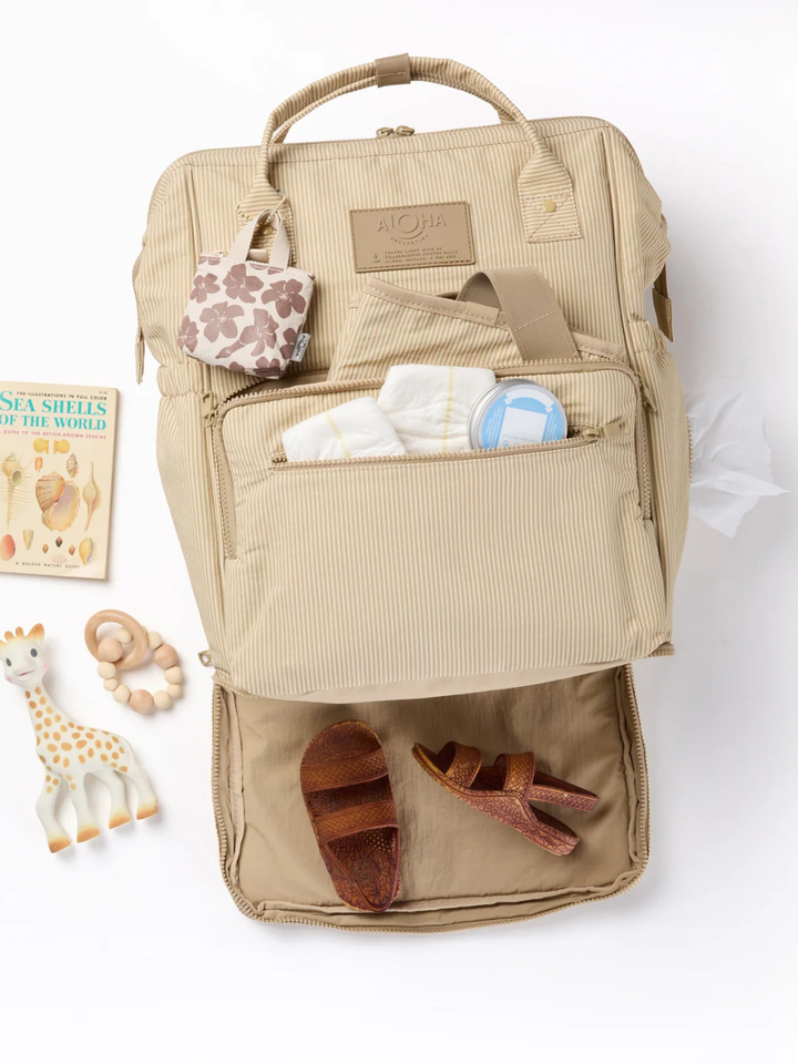 Le Bébé diaper backpack from ALOHA Collection in beige striped with front pocket, wipes, a giraffe toy, and sandals.