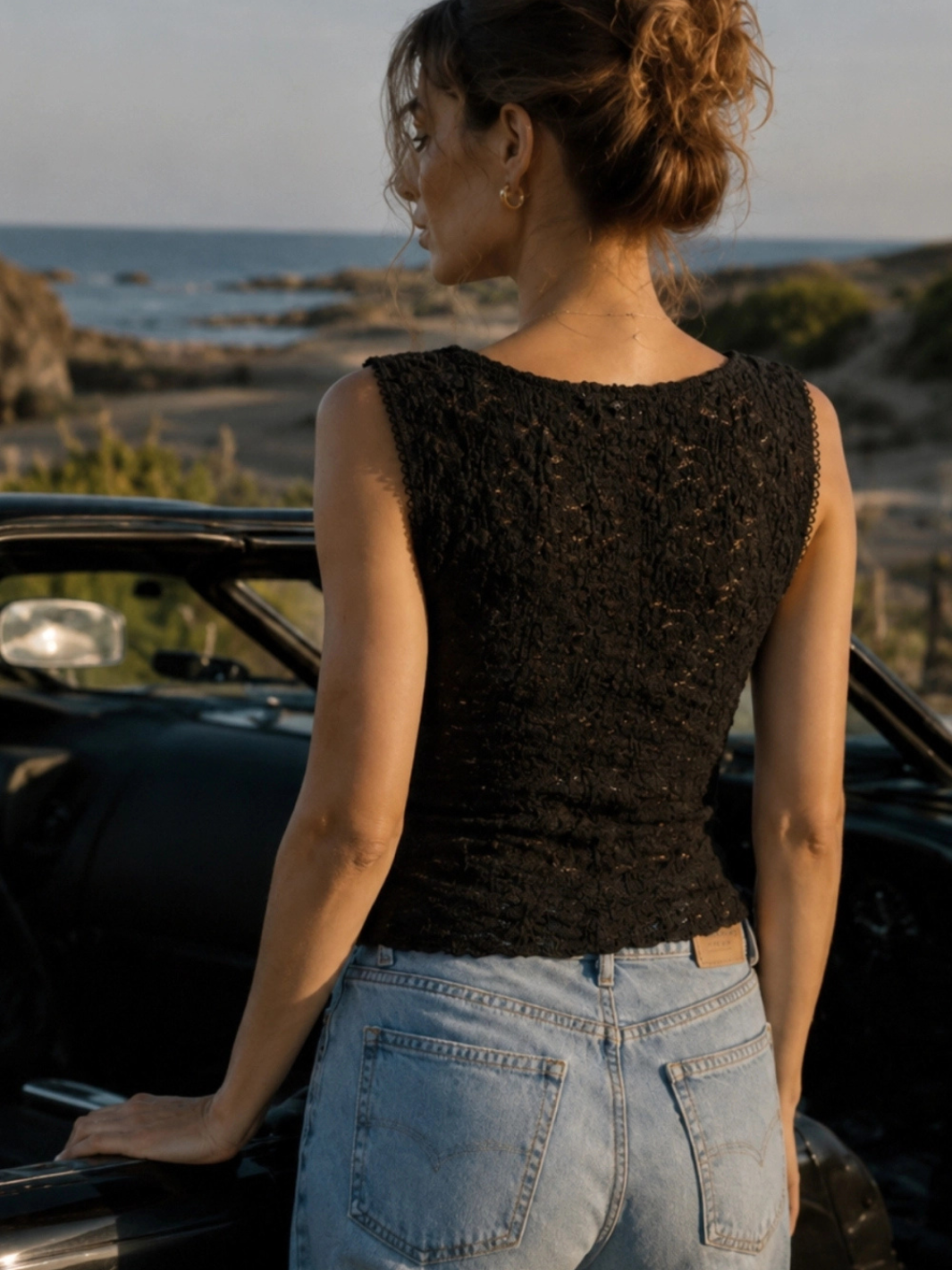 The Noir Floral Lace Cropped Tank Top in black lace, semi-sheer, worn by a woman in jeans beside a coastal car.