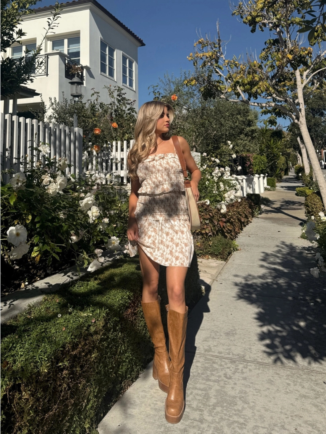 Woman in Wildflower Smocked Ruffle Mini Dress with tan knee-high boots, strolling along a sunny garden path.