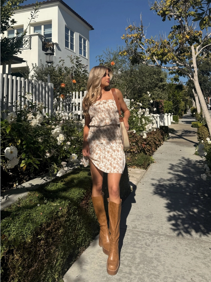 Woman in Wildflower Smocked Ruffle Mini Dress with tan knee-high boots, strolling along a sunny garden path.