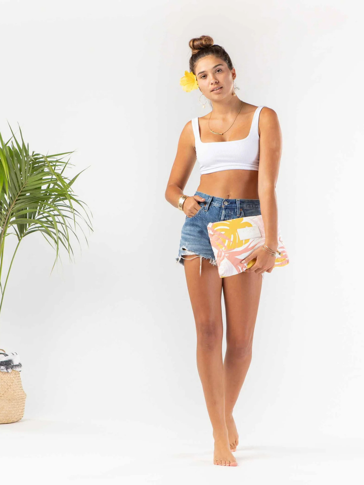 Model in ALOHA COLLECTION Tropics Starburst Tropics with white crop top, denim shorts, tropical clutch, yellow flower.