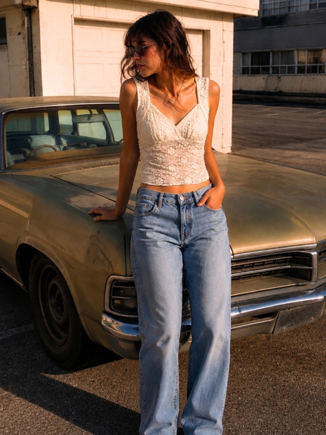 The Noir Floral Lace Cropped Tank Top on a model in white lace, styled with high-waisted jeans beside a vintage car.