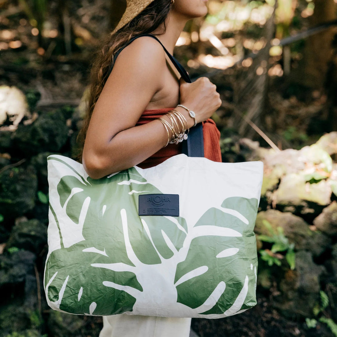 Aloha Collective Monstera Seaweed tote bag with green monstera leaf print carried by a woman outdoors.
