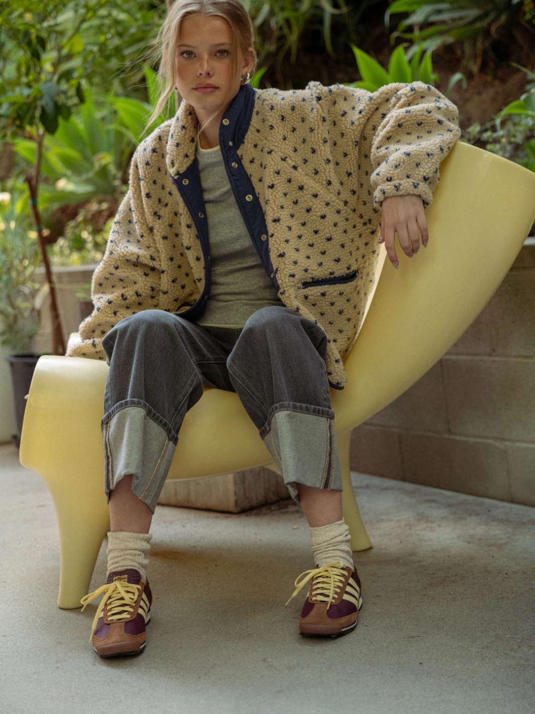 Model on a yellow curved chair wearing Canyon Bloom patterned sherpa fleece jacket with navy trim.