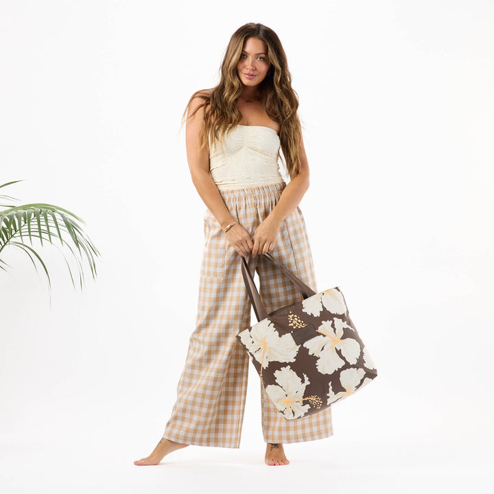 Woman in plaid trousers holding the Tahaa Day Tripper tote in coconut with cream hibiscus print