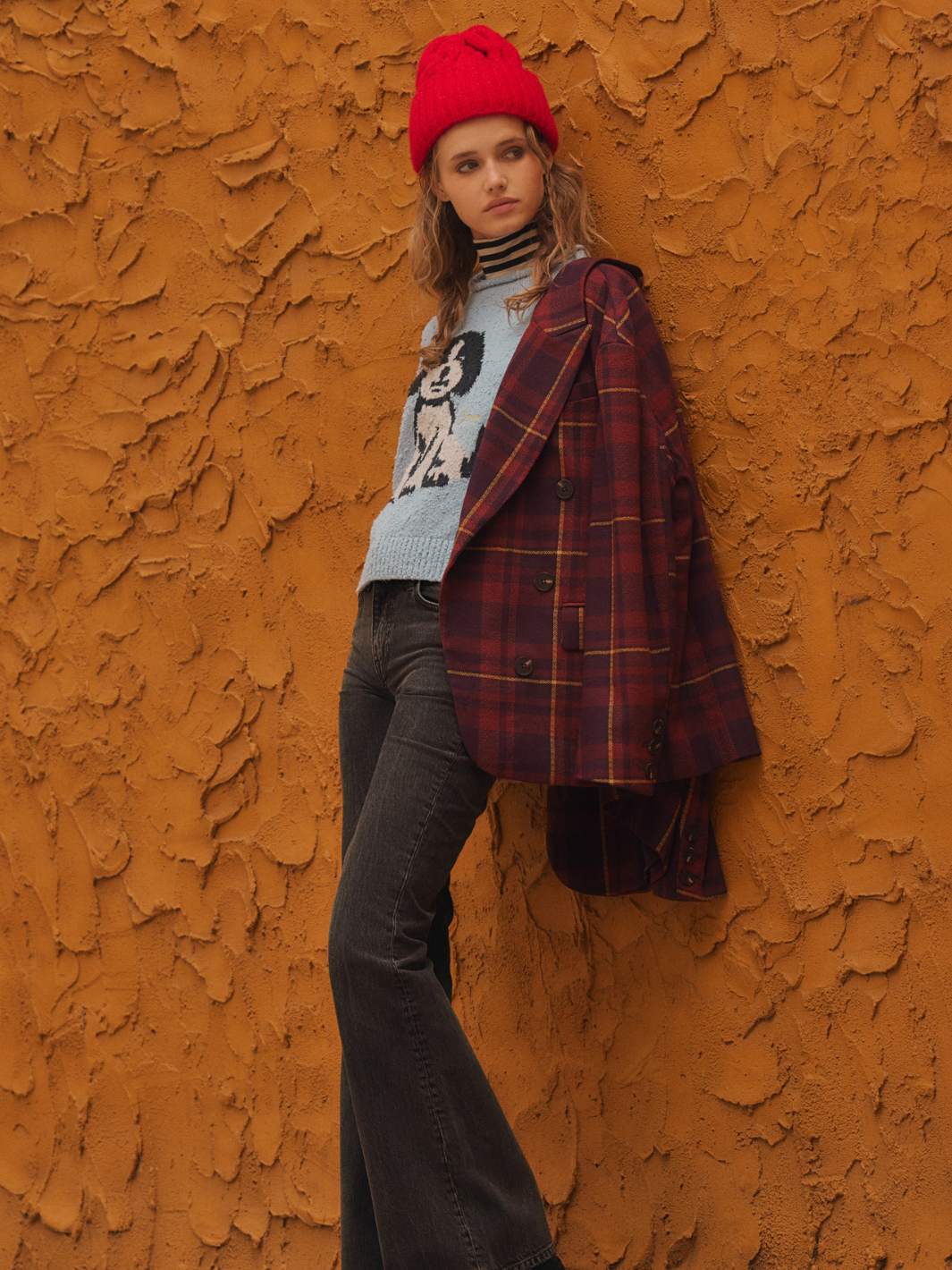Model wearing red Coast Line Beanie with chunky knit cuff, plaid coat, against textured orange wall.
