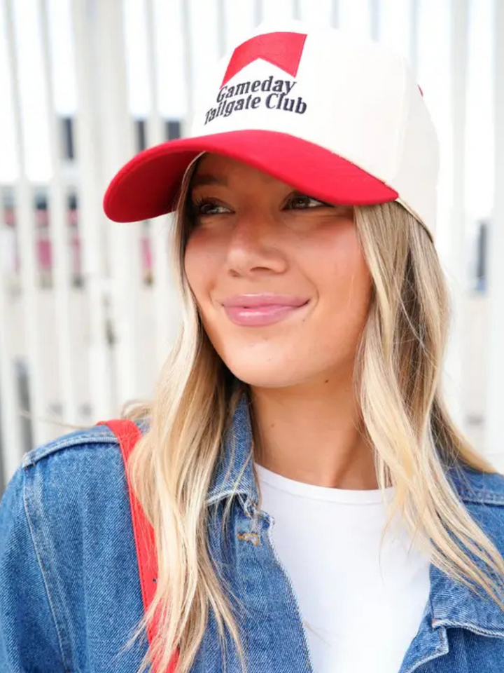 Smiling woman wearing red-and-cream Gameday Tailgate Club trucker hat with embroidered logo.
