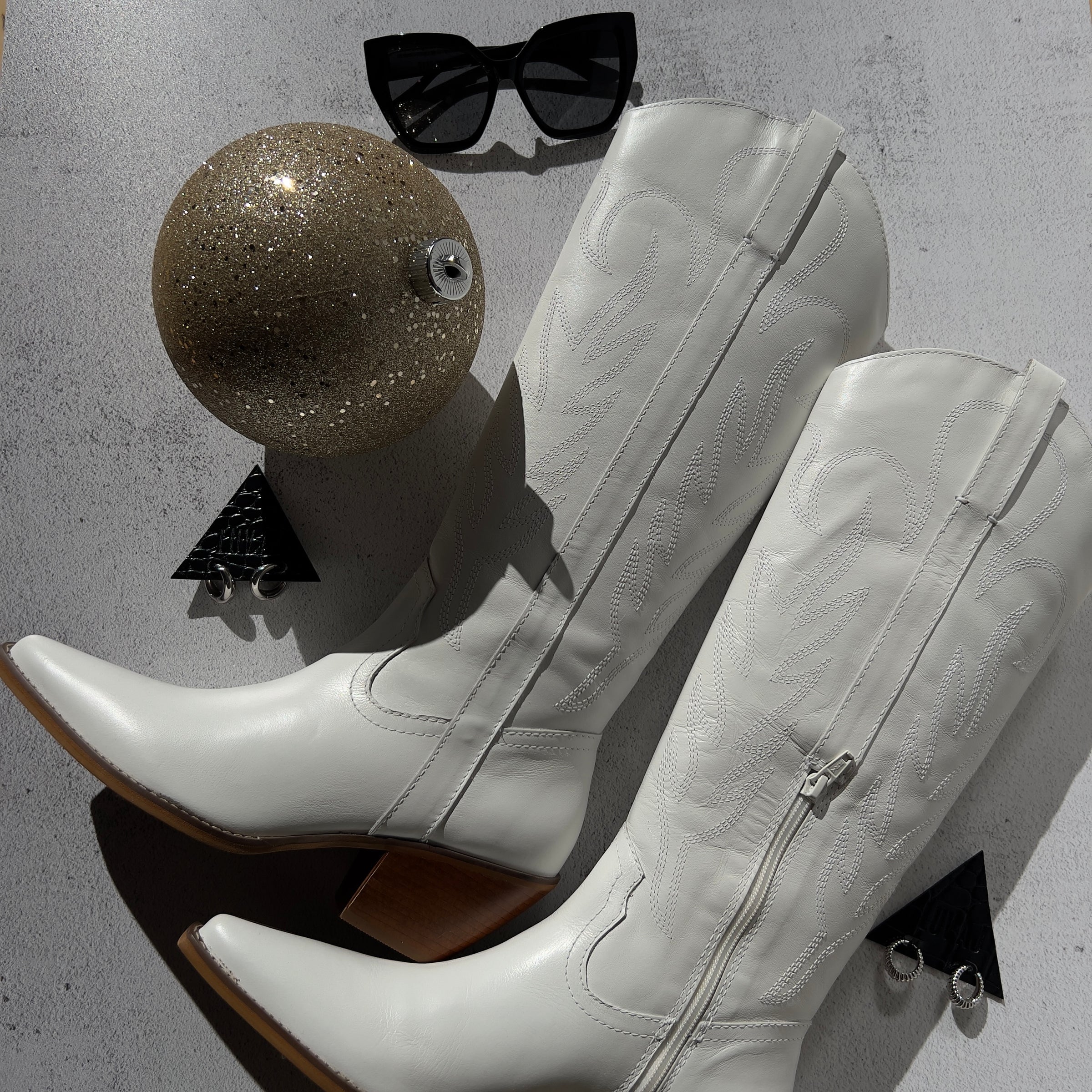 White knee-high boots with zippers, sunglasses, and a glittery ball on a gray surface