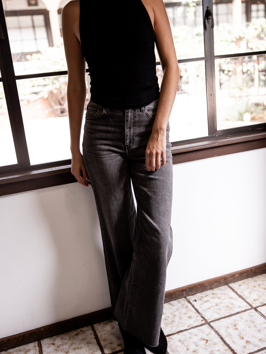 Person wearing black top and gray wide-leg jeans standing indoors with a window in the background.