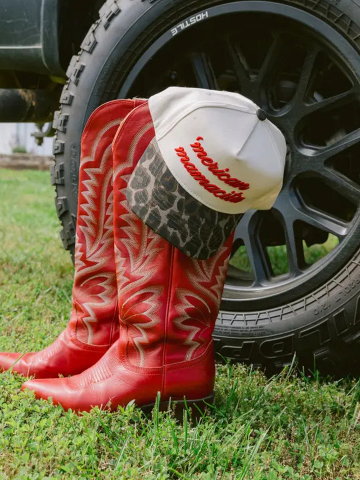 KENZKUSTOMZ - 'Merican Mamacita trucker hat paired with vibrant red cowboy boots against a tire backdrop.