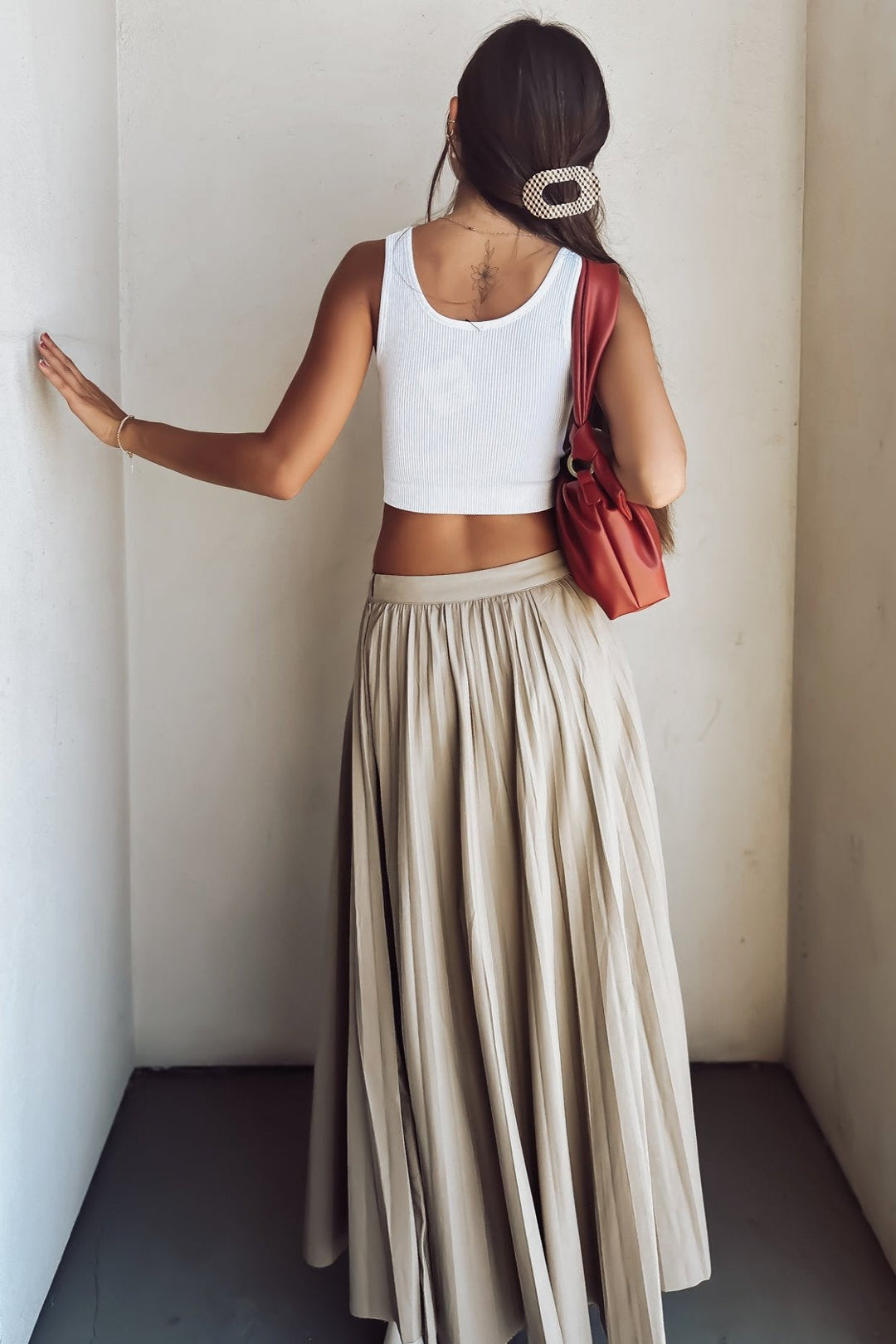 Woman in white crop top and beige pleated skirt with red shoulder bag standing against a wall.