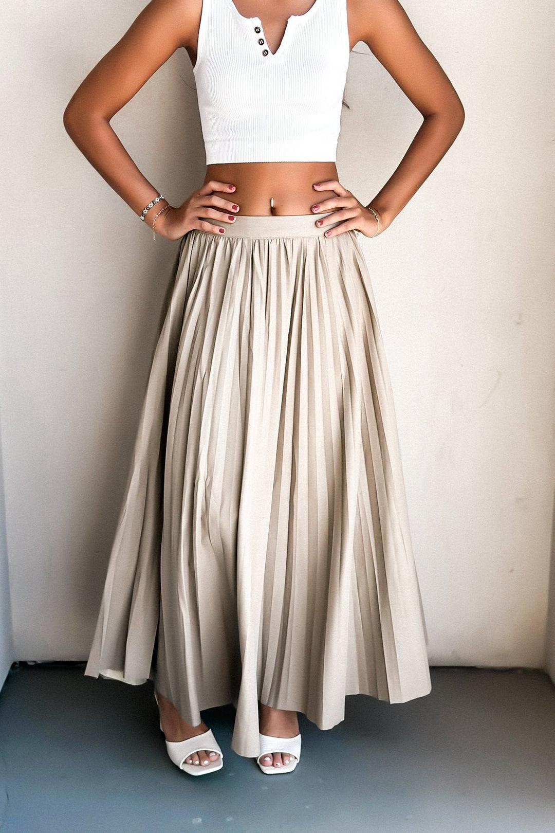 Woman in white crop top and beige pleated skirt with red shoulder bag standing against a wall.