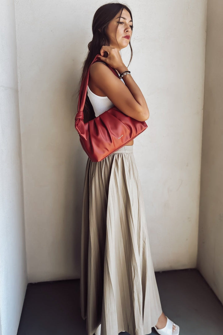 Woman in white crop top and beige pleated skirt with red shoulder bag standing against a wall.