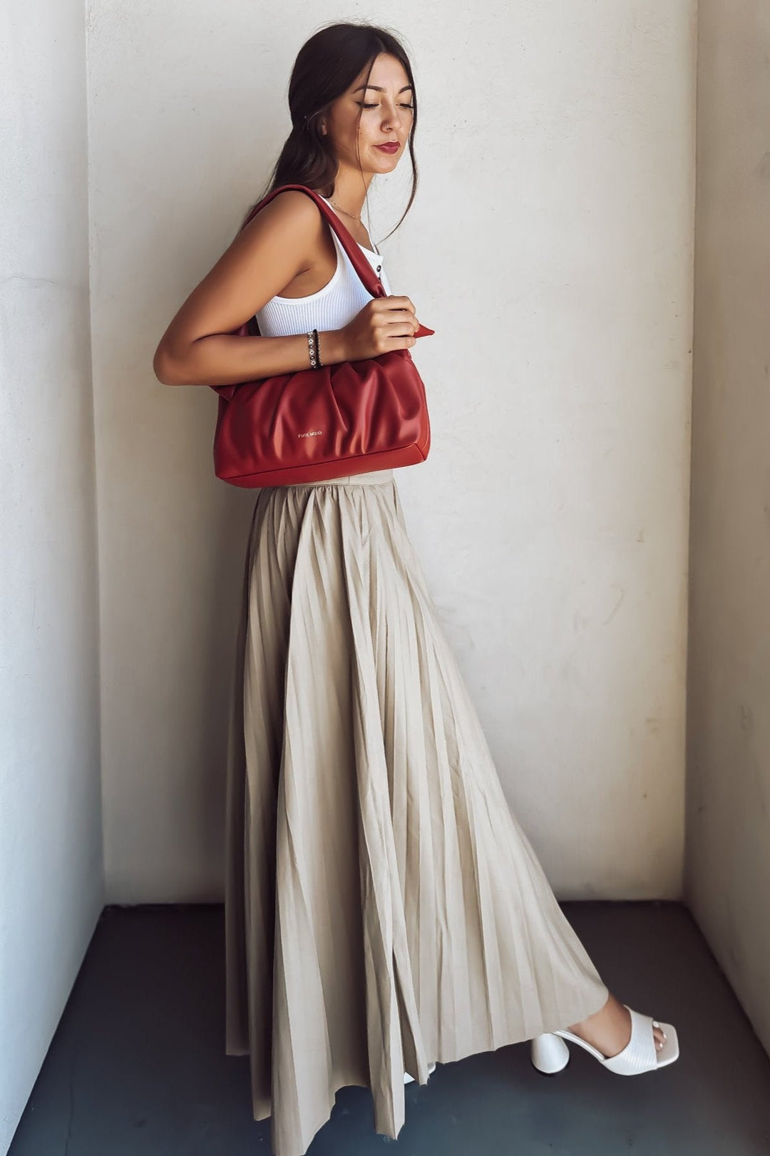 Woman in white crop top and beige pleated skirt with red shoulder bag standing against a wall.