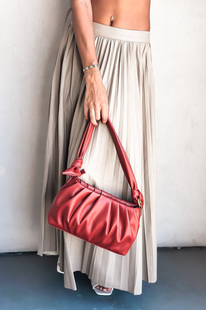 Woman in white crop top and beige pleated skirt with red shoulder bag standing against a wall.