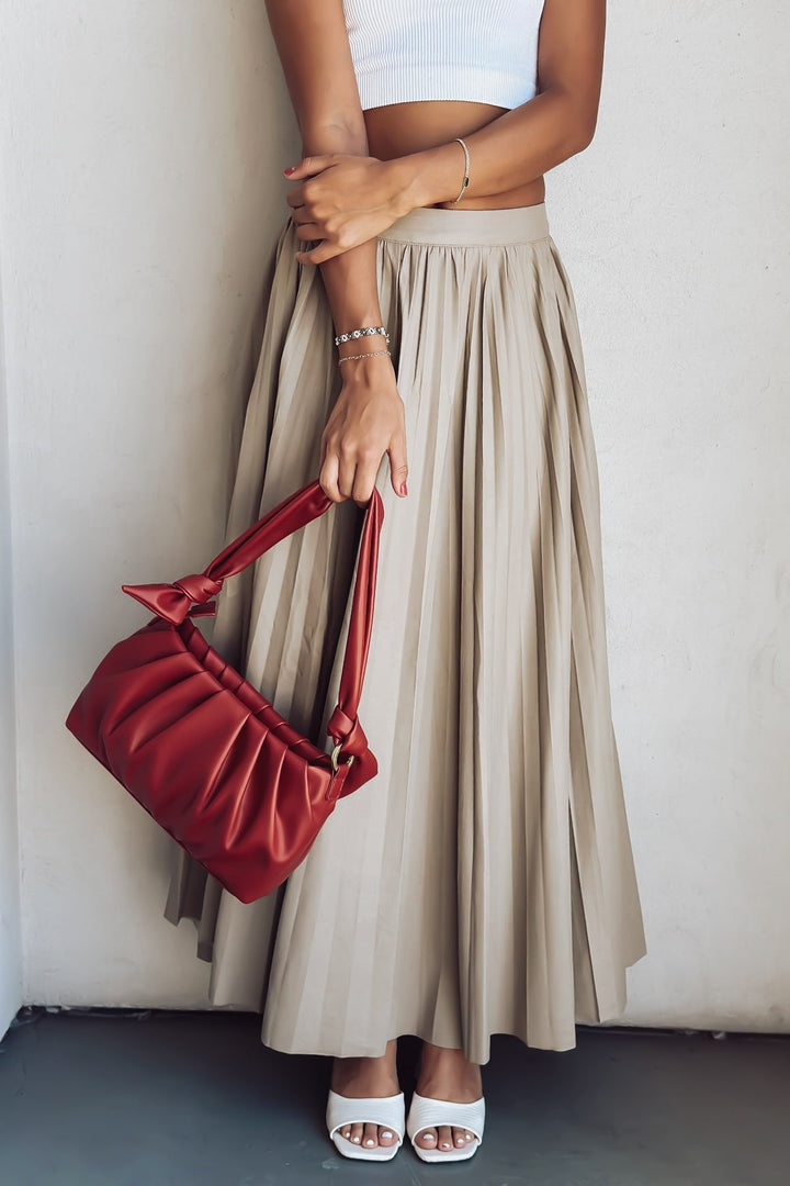 Woman in white crop top and beige pleated skirt with red shoulder bag standing against a wall.