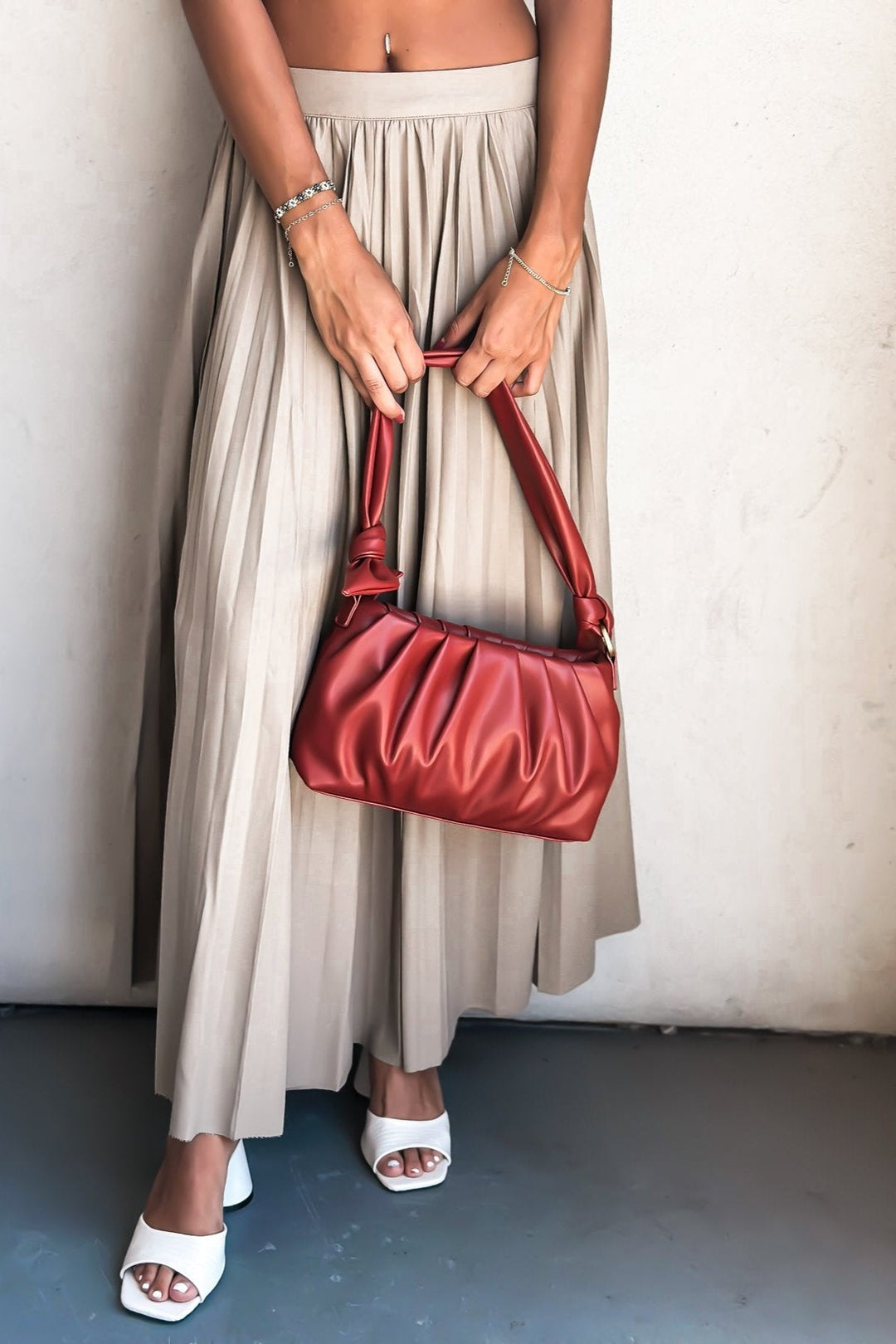 Woman in white crop top and beige pleated skirt with red shoulder bag standing against a wall.