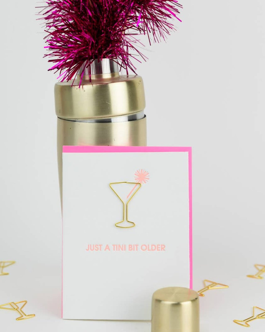 A Chez Gagne letterpress greeting card, adorned with a gold martini glass design and the phrase "Just a tini bit older" in pink text, sits beside a cocktail shaker with pink decoration on top. Martini glass paper clips from the keepsake set are scattered around, making this an ideal gift for someone special on any occasion.