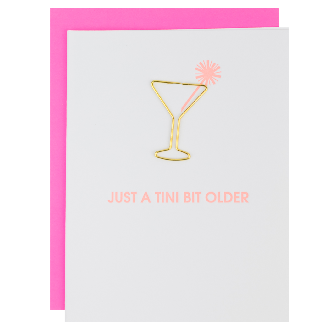 A Chez Gagne letterpress greeting card, adorned with a gold martini glass design and the phrase "Just a tini bit older" in pink text, sits beside a cocktail shaker with pink decoration on top. Martini glass paper clips from the keepsake set are scattered around, making this an ideal gift for someone special on any occasion.