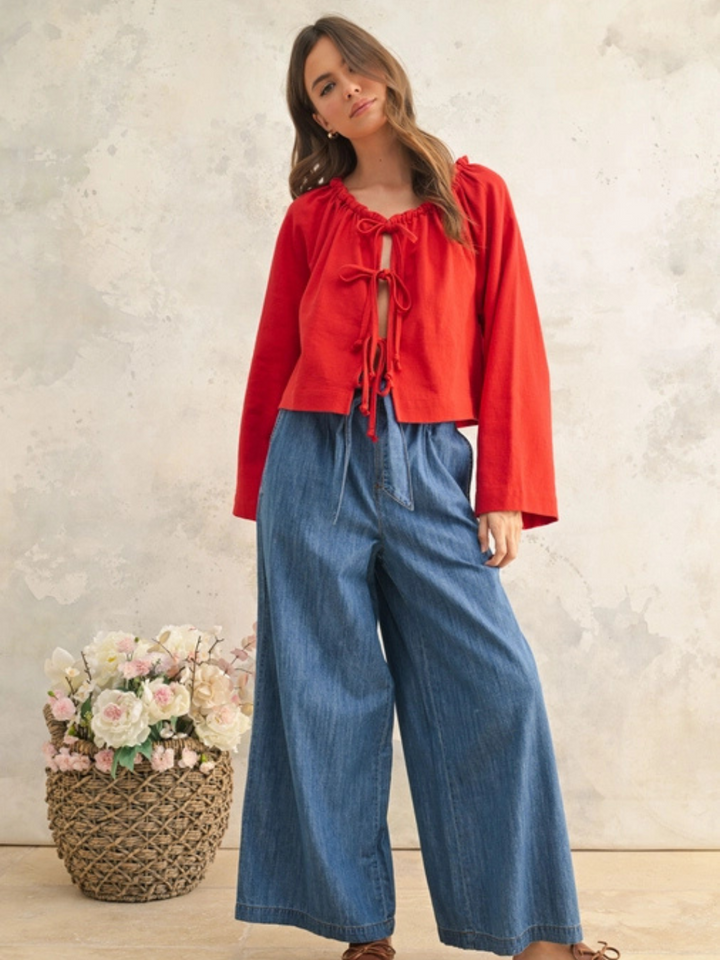 Woman posing against a neutral textured wall, wearing a red tie-front blouse with long sleeves and wide-leg denim pants. She has long, wavy brown hair and a relaxed expression. The outfit is styled with brown leather loafers, creating a casual yet chic look. A woven basket filled with pastel flowers sits beside her, adding a soft, feminine touch to the aesthetic.