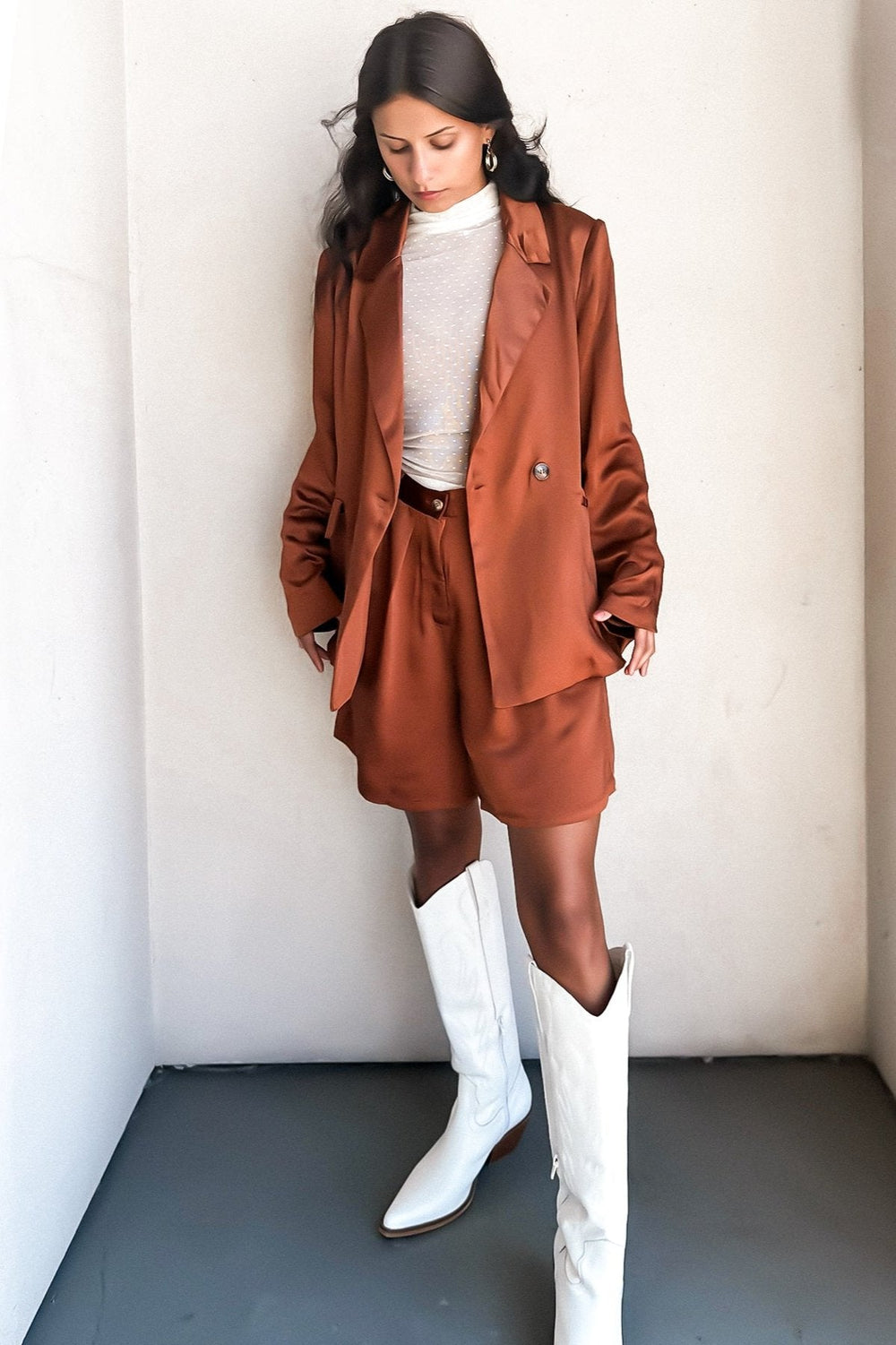 Woman in a bronze-colored bermuda short and a white top, posing against a white wall.