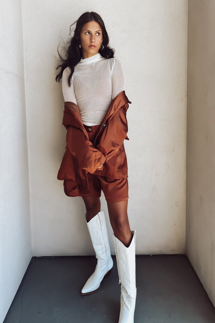 Woman in a bronze-colored bermuda short and a white top, under a matching blazer posing against a white wall.