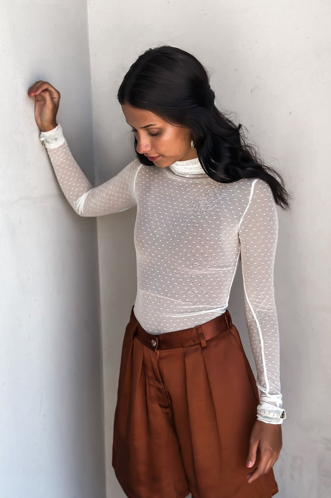 Woman in a bronze-colored bermuda short over a white top, posing against a white wall.