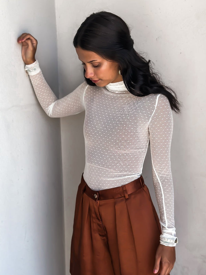 Woman in a bronze-colored bermuda short over a white top, posing against a white wall.