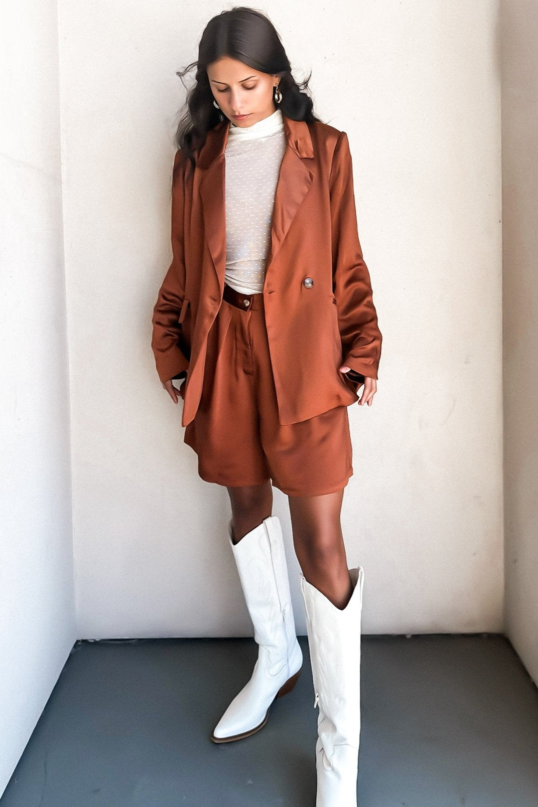 Woman in a bronze-colored blazer over a white top, posing against a white wall.