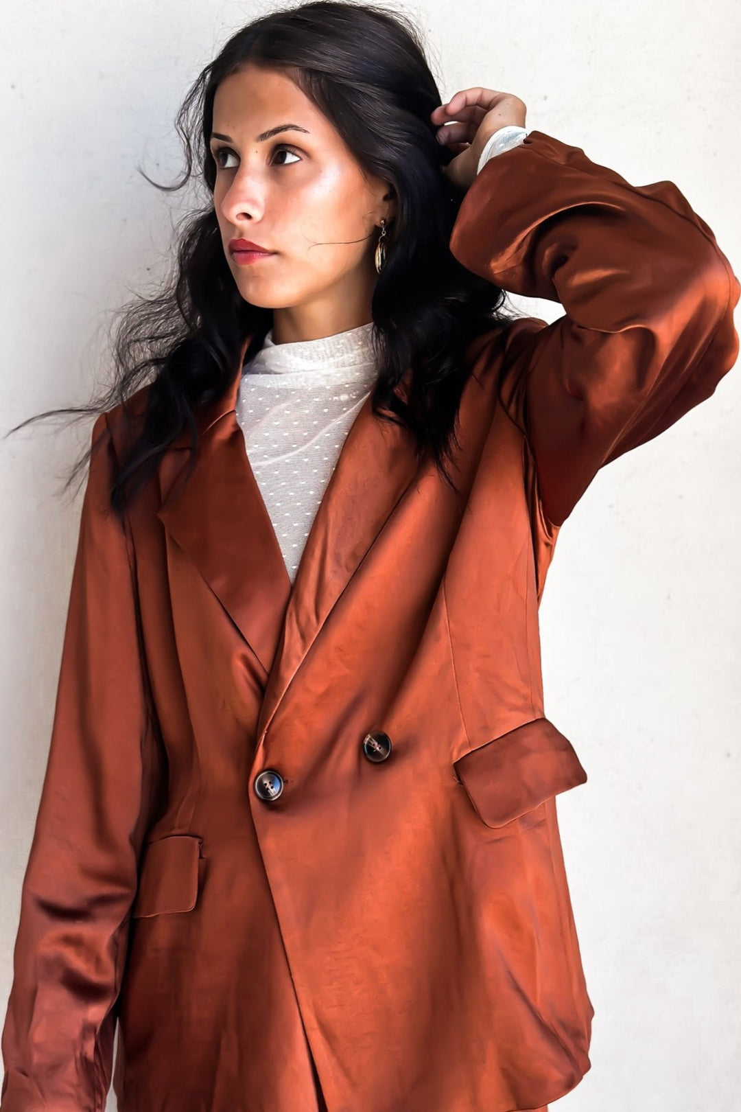 Woman in a bronze-colored blazer over a white top, posing against a white wall.