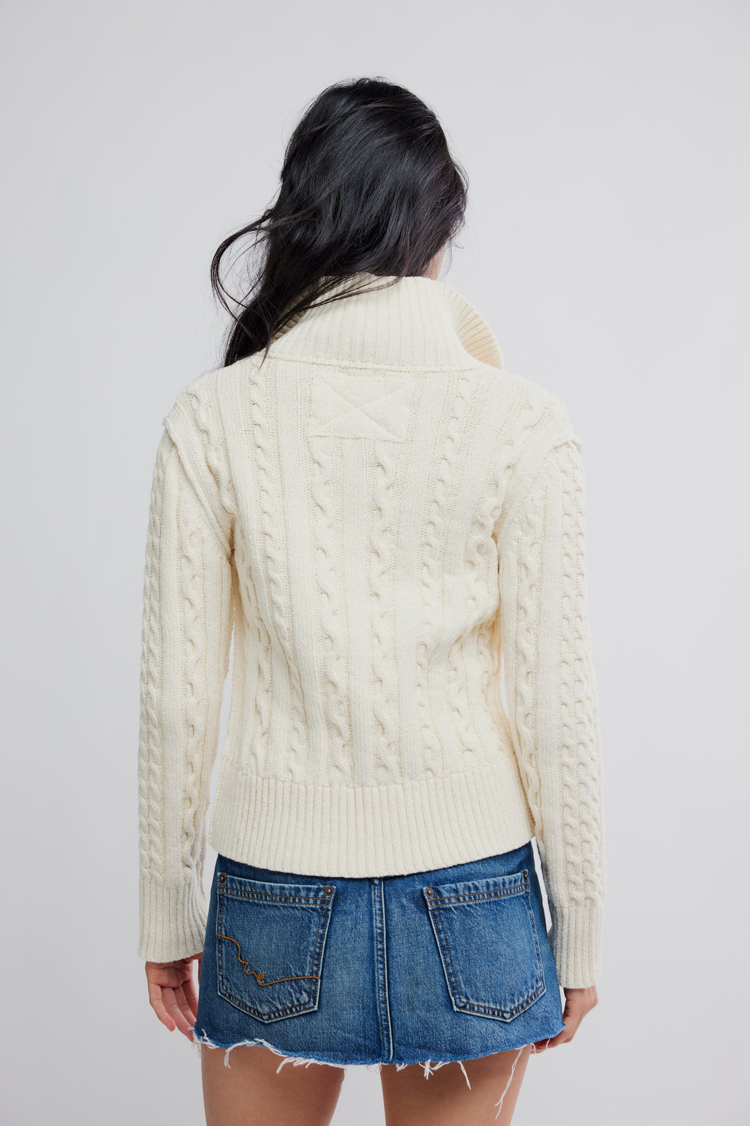 WTF Cable Collar Cardi in cream with chunky cables and a tall collar, paired with a denim mini skirt (back view).