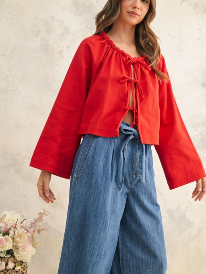 Woman wearing a bold red tie-front blouse with long bell sleeves, paired with high-waisted wide-leg denim pants. She has soft wavy brown hair and a natural makeup look. The outfit exudes effortless style with a relaxed yet polished fit. A woven basket with pastel flowers sits nearby, enhancing the boho-chic aesthetic.