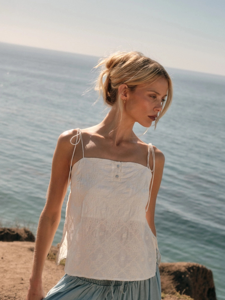 Woman wearing The Seafoam Embroidered Cami, white lace with delicate floral embroidery and spaghetti straps, by sea.