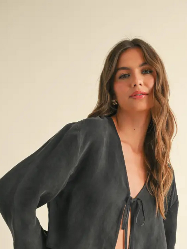 Close-up of a woman wearing a black tie-front blouse  with long sleeves, featuring a deep V-neckline. Her wavy brown hair cascades over her shoulder, and she has a soft glam makeup look with a glossy lip. The minimal backdrop enhances the chic and effortless aesthetic of the outfit.