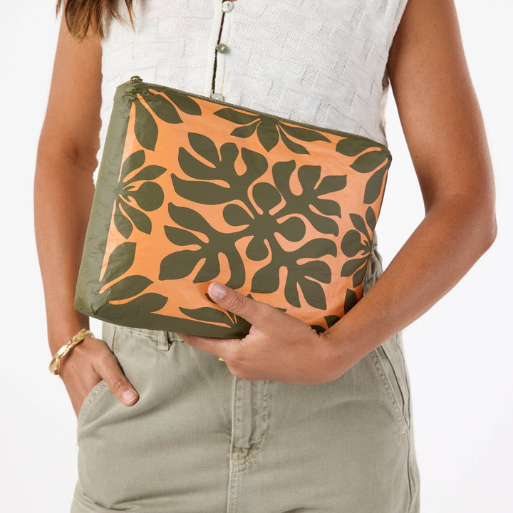 Hand holding the Kaulu Mid Pouch in papaya on olive against olive trousers, showing the ulu leaf print