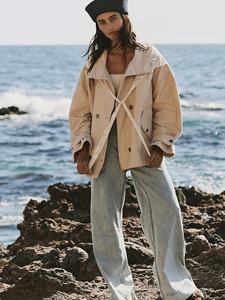 Model by the sea wearing We The Free Breezy Denim Pull-On Jeans with a beige jacket.