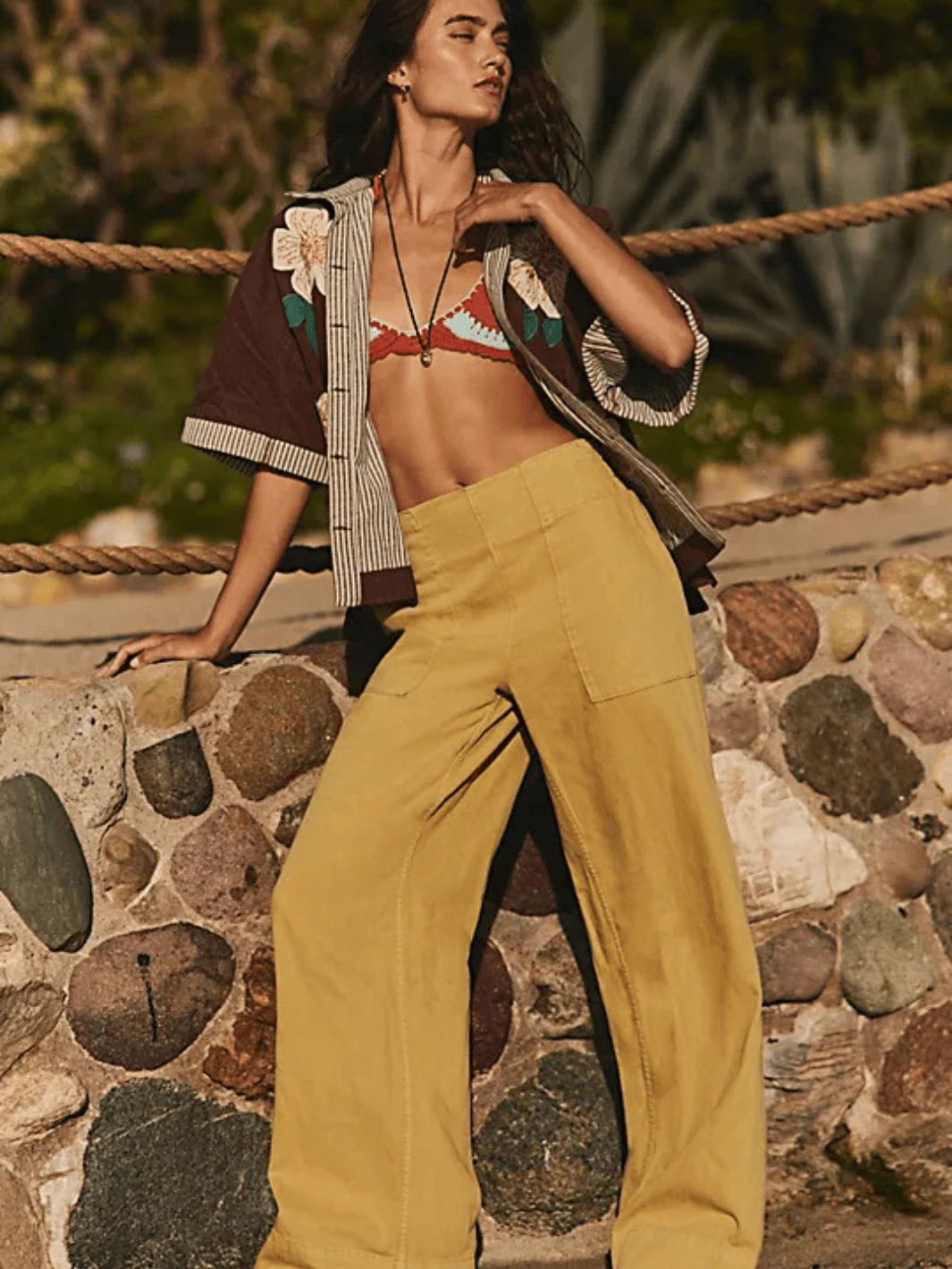 Model wearing mustard wide-leg cargo pants and bikini top outdoors by a stone wall.