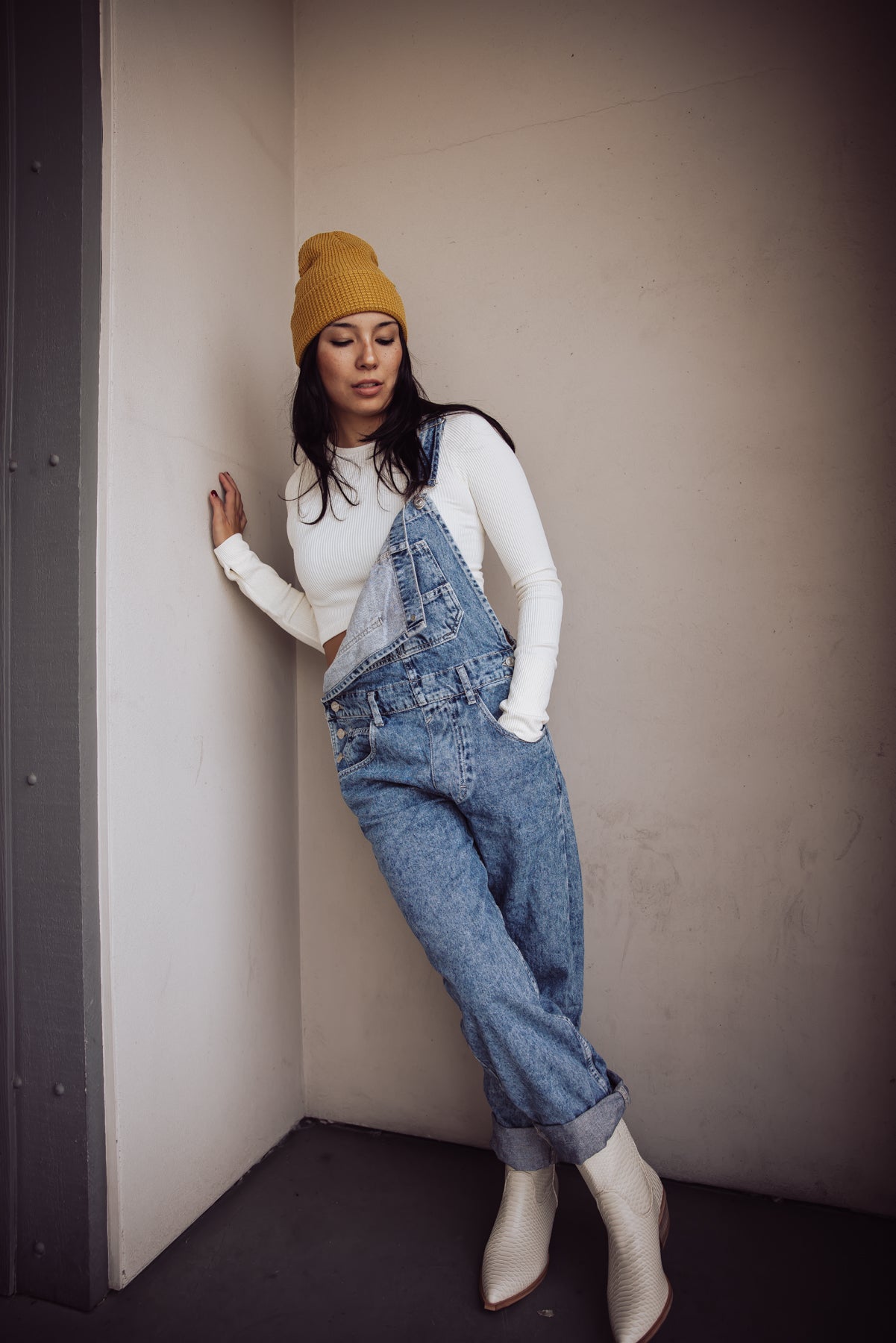 Woman styling relaxed blue denim overalls with a white long-sleeve top, mustard beanie, and ivory western boots