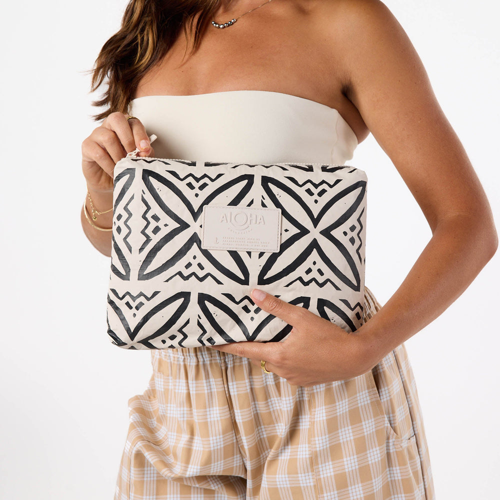 Woman in a cream top and plaid trousers holding the black-and-sandstone Pasifika Mid Pouch