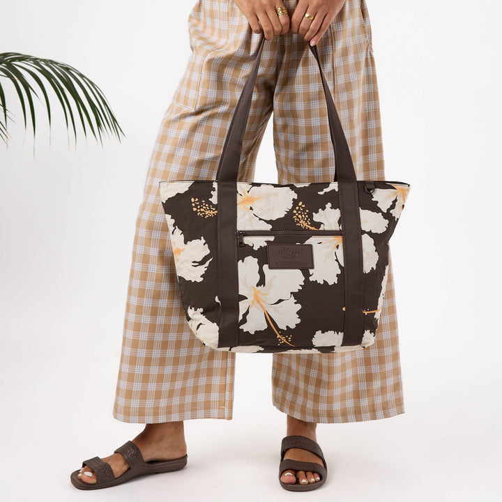 Woman in plaid trousers carrying the brown hibiscus-print Keep It Light zipper tote by its straps