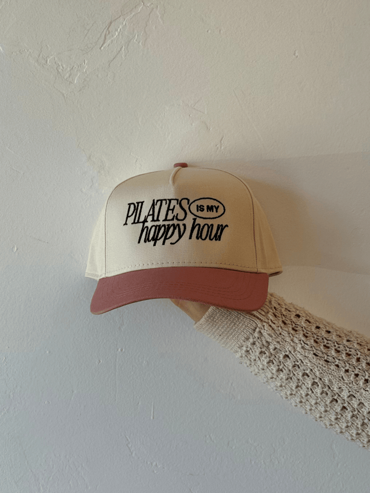 Beige Pilates Is My Happy Hour trucker hat with pink brim and black embroidered slogan.
