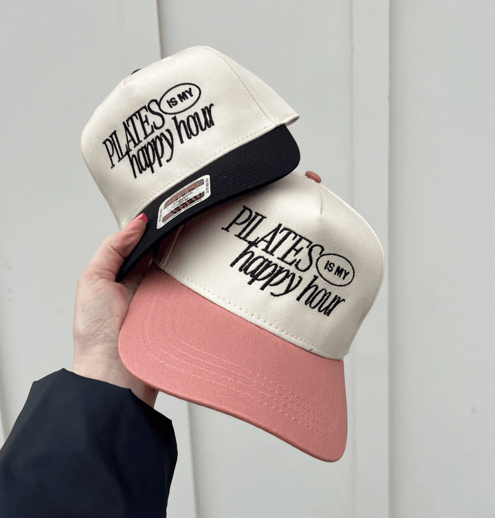 Two embroidered Pilates Is My Happy Hour trucker hats, beige fronts with black text; one pink brim and one black brim.