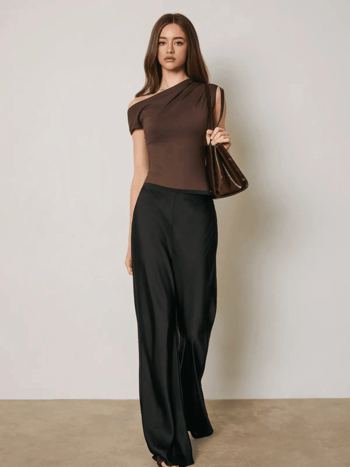 Woman wearing The After Hours Satin Straight Leg Pants in black with a high-rise, bias-cut fit and relaxed legs paired with a one-shoulder brown top and brown handbag.